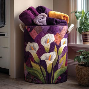 May include: A cylindrical fabric laundry hamper featuring a quilted purple patchwork design with a prominent appliqué of white calla lilies with yellow centers and green leaves. The hamper has two cream-colored side handles and is filled with folded towels.