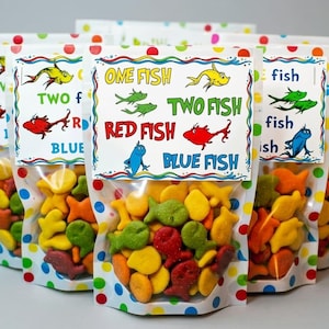May include: Clear cellophane bags filled with colorful fish-shaped crackers. Each bag has a white label with the text "One Fish, Two Fish, Red Fish, Blue Fish" and illustrations of fish. The bags are decorated with polka dots.