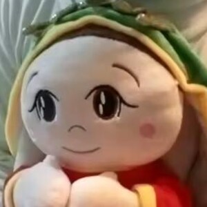 May include: A plush doll with a round face, large eyes, and a small pink cheek. The doll wears a red outfit with yellow trim and a green and yellow hat. The doll is being held.