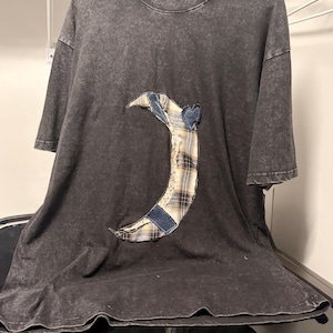 May include: A charcoal gray acid-wash t-shirt featuring a large crescent moon applique on the front. The moon is constructed from a patchwork of blue denim, beige and navy plaid fabric, and a small heart-shaped piece of blue denim at the top.