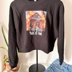 May include: Black sweatshirt with a graphic of a woman in sunglasses in front of a burning house. The text "I'm fine, this is fine" is below the image. The sweatshirt has long sleeves.