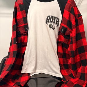 May include: A long-sleeve shirt featuring a white center panel with a black graphic of dice and the text ADTR. The sleeves and chest pockets are made of a red and black buffalo plaid fabric, while the collar and shoulder panels are solid black.