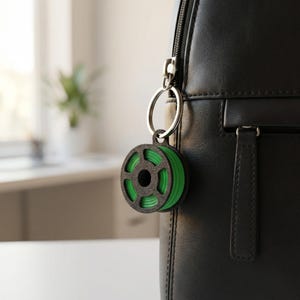 Könnte beinhalten: Ein schwarzer und grüner 3D-Drucker-Filament-Spulen-Schlüsselanhänger, der an einem schwarzen Rucksack hängt. Der Schlüsselanhänger hat ein kreisförmiges Design mit einer grünen Filamentrolle und einem schwarzen Rahmen. Der Schlüsselanhänger ist an einem silbernen Schlüsselring befestigt.