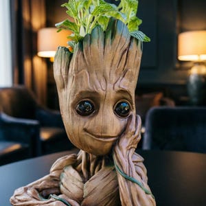 May include: A brown Groot-shaped planter with green plants. The planter has a smiling face and is resting its chin on its hand. The planter is made of a wood-like material and has a detailed texture. The plant has green leaves with white veins.