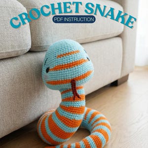 Puede incluir: Un juguete de serpiente de ganchillo a rayas azules y naranjas, con una lengua roja. El texto "CROCHET SNAKE PDF INSTRUCTION" está encima. La serpiente está sobre un suelo de madera, junto a un sofá beige.