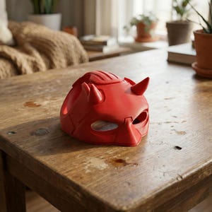 K&ouml;nnte beinhalten: Eine rote Maske mit H&ouml;rnern und Augenaussparungen liegt auf einem Holztisch. Die Maske hat eine strukturierte Oberfl&auml;che und ein Design, das an eine Superheldenmaske erinnert.