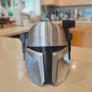 Puede incluir: Un casco mandaloriano plateado y negro, con una visera en forma de T y un dise&ntilde;o angular. El casco tiene una superficie texturizada y probablemente est&aacute; impreso en 3D. Incluye un accesorio negro en el lateral y dos peque&ntilde;os tri&aacute;ngulos en la parte superior.