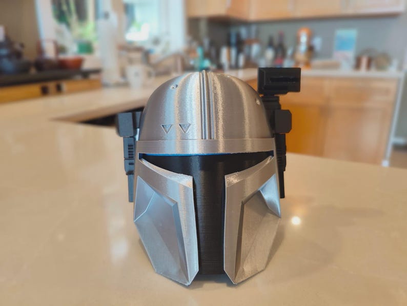 Puede incluir: Un casco mandaloriano plateado y negro, con una visera en forma de T y un dise&ntilde;o angular. El casco tiene una superficie texturizada y probablemente est&aacute; impreso en 3D. Incluye un accesorio negro en el lateral y dos peque&ntilde;os tri&aacute;ngulos en la parte superior.
