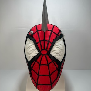 May include: A red and black Spider-Man mask with a silver spike on top. The mask features white mesh eye openings and a black web design. A silver ring and silver studs are on the mask.