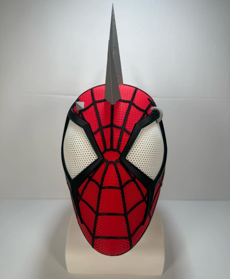 May include: A red and black Spider-Man mask with a silver spike on top. The mask features white mesh eye openings and a black web design. A silver ring and silver studs are on the mask.