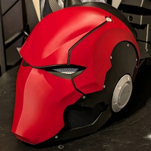 May include: A red and black helmet with a futuristic design. The helmet has a textured red surface with black accents, including a mesh area near the mouth and a circular silver detail on the side. The helmet has a sleek, angular shape.