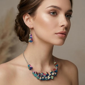 Puede incluir: Una mujer con un collar geométrico colorido y pendientes a juego. La joyería presenta pequeñas cuentas en forma de cubo en tonos de azul, rosa, verde y morado. El collar es de cadena de plata.