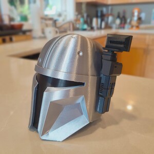 Puede incluir: Un casco mandaloriano plateado y negro, una r&eacute;plica de la franquicia Star Wars. El casco presenta una visera en forma de T, un tel&eacute;metro y paneles detallados. El casco est&aacute; hecho de un material met&aacute;lico y se encuentra sobre una superficie clara.