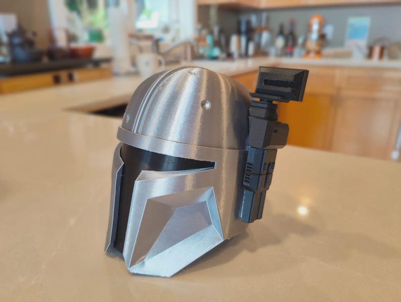 Puede incluir: Un casco mandaloriano plateado y negro, una r&eacute;plica de la franquicia Star Wars. El casco presenta una visera en forma de T, un tel&eacute;metro y paneles detallados. El casco est&aacute; hecho de un material met&aacute;lico y se encuentra sobre una superficie clara.