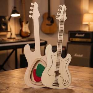 Può includere: Due supporti in legno a forma di chitarra. Uno è una forma di chitarra piena, e l'altro ha un ritaglio a forma di chitarra con plettri in feltro rosso e verde all'interno. Le chitarre sono in legno chiaro e poggiano su una superficie di legno.