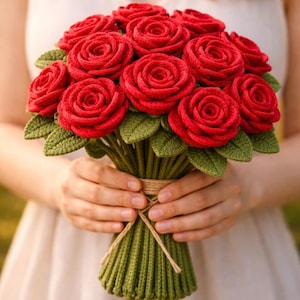 以下が含まれることがあります： 赤いかぎ針編みのバラの花束。緑の葉と茎があり、ひもで結ばれています。手作りの花は白いドレスを着た人が持っています。