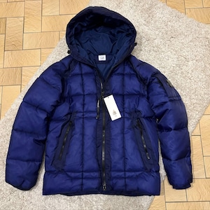 Può includere: Giacca puffer trapuntata blu navy con cappuccio. La giacca ha una cerniera a tutta lunghezza, due tasche laterali e una piccola toppa con logo sulla manica. Un'etichetta bianca pende dalla cerniera.