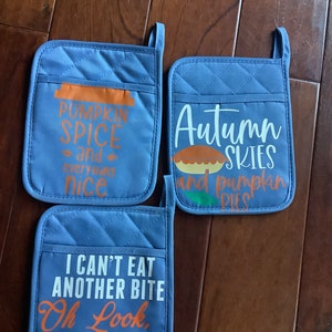 May include: Three blue quilted oven mitts with white and orange text. The mitts have phrases like "Pumpkin Spice and everything nice", "Autumn Skies and pumpkin pies", and "I can't eat another bite, Oh look, pie!"