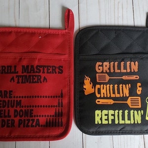 May include: Two red and black oven mitts with a grilling theme. The red mitt has the text "Grill Master's A Timer A" and a list of doneness levels for meat. The black mitt has the text "Grillin Chillin' & Refillin'" with a flame, spatula, and beer mug graphic.