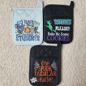Peut inclure: Trois maniques de cuisine matelass&eacute;es noires et bleues avec des motifs sur le th&egrave;me d'Halloween. Une manique a une sorci&egrave;re sur un balai avec le texte "Witch Please! Bake Me Some Cookies". Une autre manique a un cr&acirc;ne et une toile d'araign&eacute;e avec le texte "One Spook Tacular Teacher". La troisi&egrave;me manique a une citrouille et le texte "I Teach The Most Spook Tacular Students".