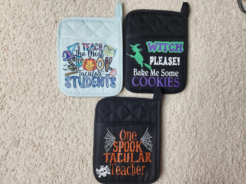 Peut inclure: Trois maniques de cuisine matelass&eacute;es noires et bleues avec des motifs sur le th&egrave;me d'Halloween. Une manique a une sorci&egrave;re sur un balai avec le texte "Witch Please! Bake Me Some Cookies". Une autre manique a un cr&acirc;ne et une toile d'araign&eacute;e avec le texte "One Spook Tacular Teacher". La troisi&egrave;me manique a une citrouille et le texte "I Teach The Most Spook Tacular Students".