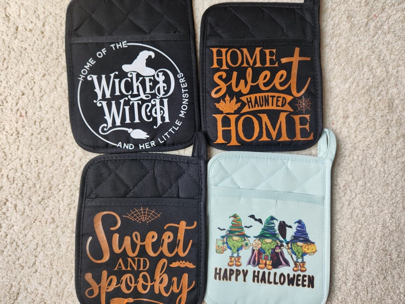 Peut inclure: Quatre maniques de four sur le th&egrave;me d'Halloween. Deux maniques noires avec du texte orange, l'une avec "Home of the Wicked Witch and her little monsters" et l'autre avec "Home sweet haunted home". Une manique bleue avec un message "Happy Halloween" et trois gnomes. Une manique noire avec du texte orange qui dit "Sweet and spooky".
