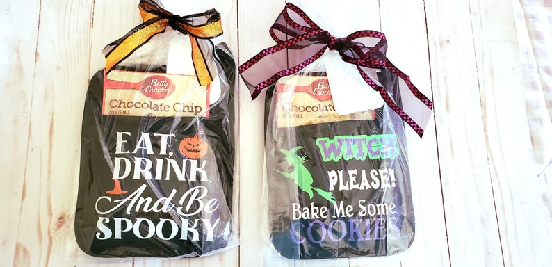 Peut inclure: Deux ensembles de torchons de cuisine sur le th&egrave;me d'Halloween, chacun emball&eacute; dans du plastique transparent avec un ruban noir. Le premier ensemble comprend un torchon noir avec du texte blanc qui dit "Eat, Drink and Be Spooky" et un graphique de citrouille. Le deuxi&egrave;me ensemble comprend un torchon noir avec du texte blanc qui dit "Witches Please! Bake Me Some Cookies" et un graphique de sorci&egrave;re verte. Les deux ensembles comprennent une bo&icirc;te de m&eacute;lange &agrave; biscuits aux p&eacute;pites de chocolat Betty Crocker.