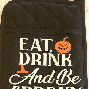 Peut inclure: Manique de four noire avec du texte blanc qui dit "Eat, Drink and Be Spooky". Une petite citrouille orange avec un visage de citrouille est &agrave; c&ocirc;t&eacute; du mot "Eat".