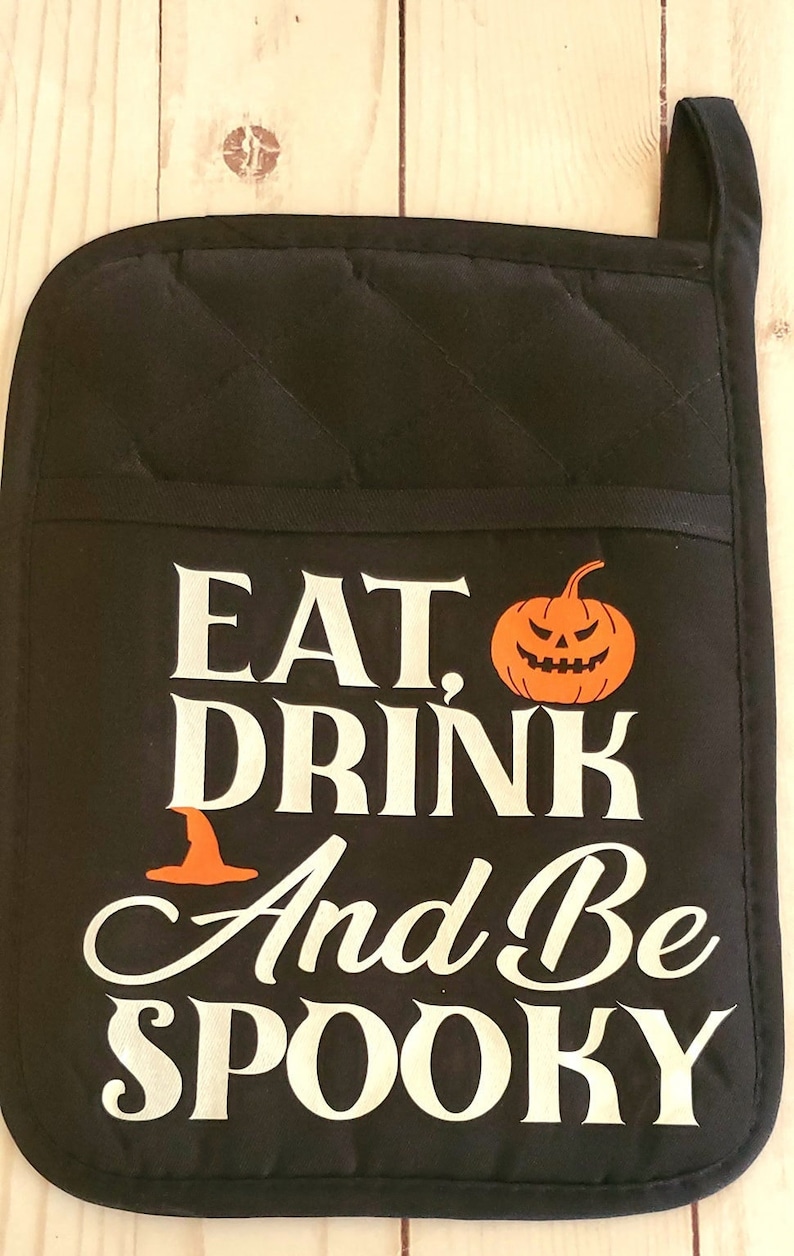 Peut inclure: Manique de four noire avec du texte blanc qui dit "Eat, Drink and Be Spooky". Une petite citrouille orange avec un visage de citrouille est &agrave; c&ocirc;t&eacute; du mot "Eat".