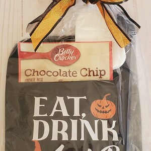 Peut inclure: Un torchon de cuisine noir et blanc sur le th&egrave;me d'Halloween avec le texte "Eat, Drink and Be Spooky" et un graphique de citrouille. Un paquet de m&eacute;lange &agrave; biscuits aux p&eacute;pites de chocolat Betty Crocker est inclus dans le sac cadeau.