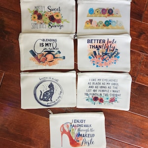 May include: Set of ten white canvas zippered pouches with various colorful designs and text. The pouches feature sayings such as "Mostly Sweet Sometimes Savage", "Blending is My Cardio", "Better Late Than Ugly", "Karma is a Cat", "I Like My Eyelashes as Black as My Soul", and "I Enjoy a Long Walk Through the Makeup Aisle".