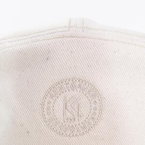 May include: Off-white baseball cap with a stitched brim and a circular logo. The logo features the letters "KI" inside a dotted circle with surrounding text. The cap is made of a textured fabric.