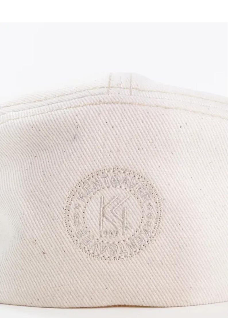 May include: Off-white baseball cap with a stitched brim and a circular logo. The logo features the letters "KI" inside a dotted circle with surrounding text. The cap is made of a textured fabric.