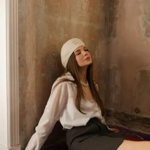May include: A woman wearing a white beret, white shirt, and dark gray skirt. She is wearing a pearl necklace and holding a white beaded clutch. The background is a textured wall with a red and brown rug.
