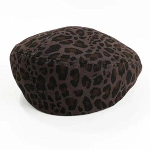 May include: A brown beret with a leopard print pattern. The hat features dark brown spots on a lighter brown background. The beret is a classic style, suitable for various outfits and occasions.