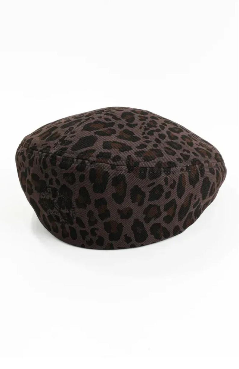 May include: A brown beret with a leopard print pattern. The hat features dark brown spots on a lighter brown background. The beret is a classic style, suitable for various outfits and occasions.