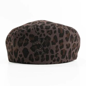 May include: A brown leopard print beret. The hat features a repeating pattern of dark brown spots on a lighter brown background. A dark emblem is centered on the front of the hat.