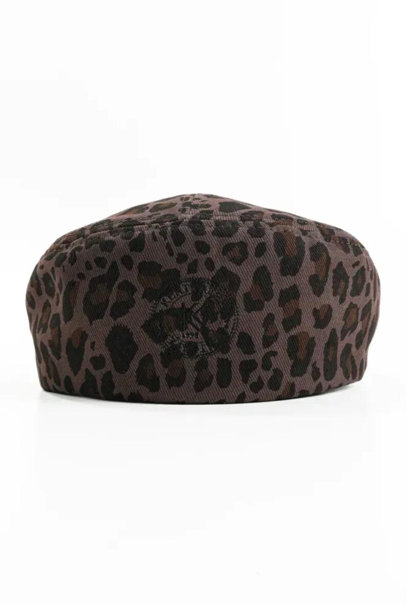 May include: A brown leopard print beret. The hat features a repeating pattern of dark brown spots on a lighter brown background. A dark emblem is centered on the front of the hat.