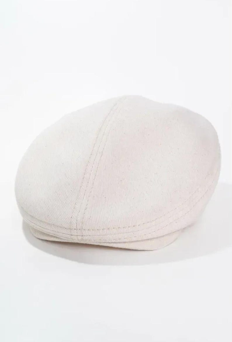 May include: A light beige newsboy cap. The hat has a rounded crown and a short, stiff brim. The cap is made of a textured fabric and features a stitched seam detail running down the center of the crown.