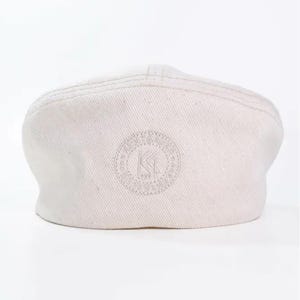 May include: Off-white, flat cap with a rounded crown and a small, curved brim. The cap features a stitched design on the crown and an embroidered circular logo with the letters "KI" in the center. The cap is made of a textured fabric.