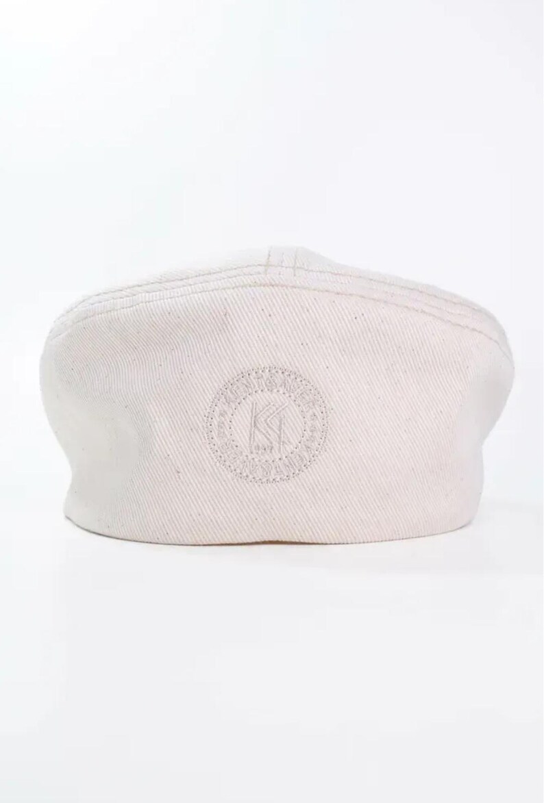 May include: Off-white, flat cap with a rounded crown and a small, curved brim. The cap features a stitched design on the crown and an embroidered circular logo with the letters "KI" in the center. The cap is made of a textured fabric.