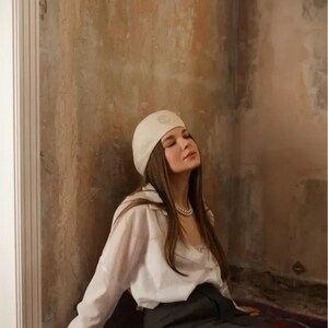 May include: A woman wearing a white beret, white button-down shirt, and a dark gray skirt. She is seated on a purple rug, with a small white beaded purse. The background is a textured wall.