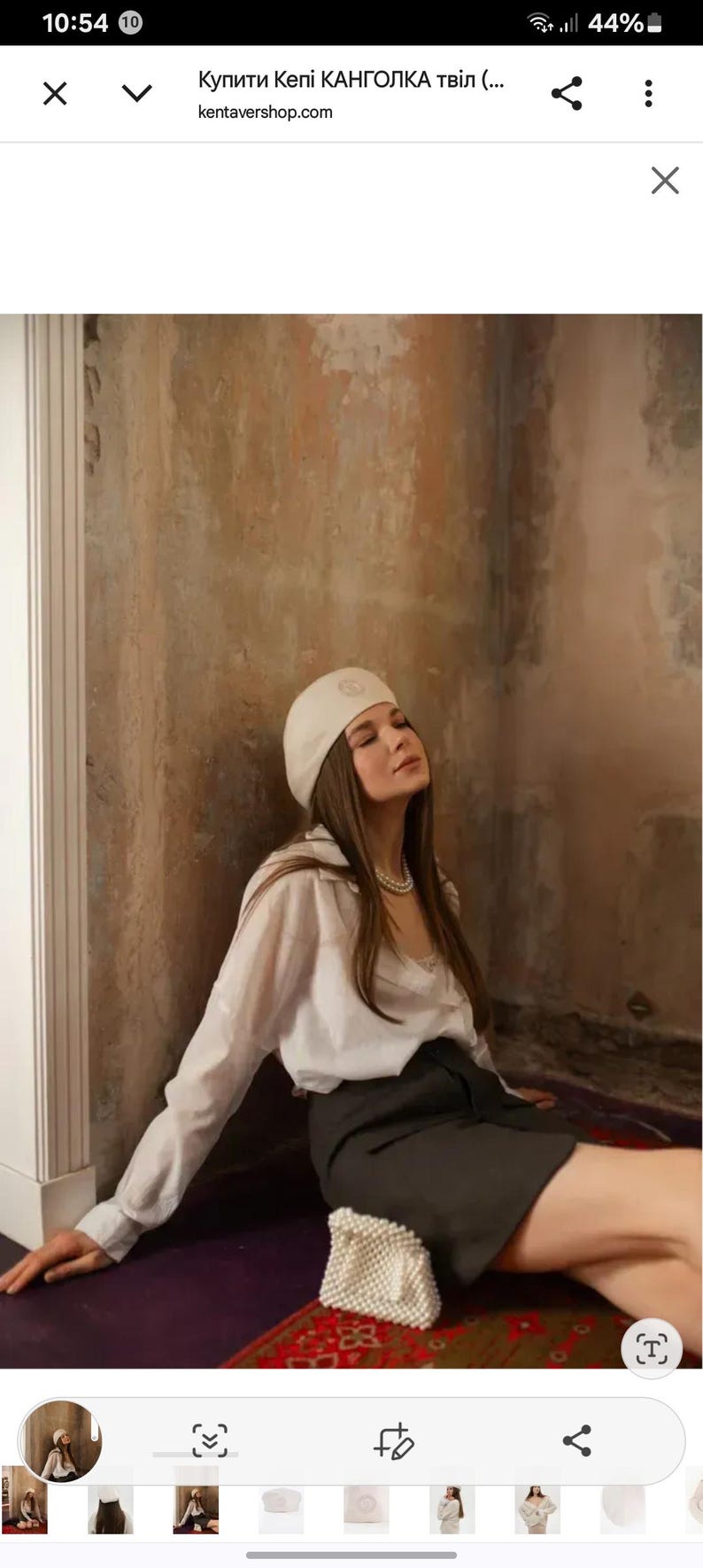 May include: A woman wearing a white beret, white button-down shirt, and a dark gray skirt. She is seated on a purple rug, with a small white beaded purse. The background is a textured wall.