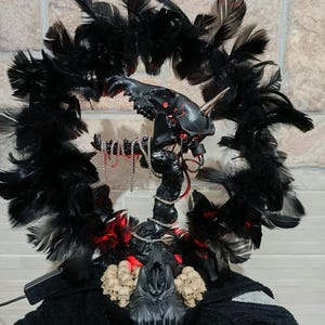 May include: A gothic-themed sculpture featuring a black, horned creature with red accents, surrounded by a wreath of black feathers. The base includes a grim reaper figure and several skulls. The sculpture has a dark, ominous aesthetic.