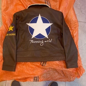 K&ouml;nnte beinhalten: Eine braune Jacke mit cremefarbenem Kragen, die auf der R&uuml;ckseite einen gro&szlig;en wei&szlig;en Stern in einem blauen Kreis zeigt. Unter dem Stern ist der Text Running wild aufgedruckt. Ein gelber Stern und der Text AIR BORNE befinden sich am rechten &Auml;rmel.