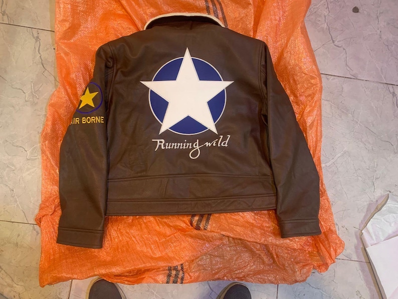 K&ouml;nnte beinhalten: Eine braune Jacke mit cremefarbenem Kragen, die auf der R&uuml;ckseite einen gro&szlig;en wei&szlig;en Stern in einem blauen Kreis zeigt. Unter dem Stern ist der Text Running wild aufgedruckt. Ein gelber Stern und der Text AIR BORNE befinden sich am rechten &Auml;rmel.