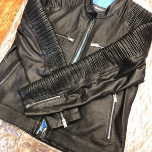 K&ouml;nnte beinhalten: Eine schwarze Lederjacke mit Stehkragen, leuchtend blauem Innenfutter und silberfarbenen Metallrei&szlig;verschl&uuml;ssen. Die &Auml;rmel und Schultern sind mit horizontalen Rippeneins&auml;tzen versehen und der Text MADE IN ist sichtbar.