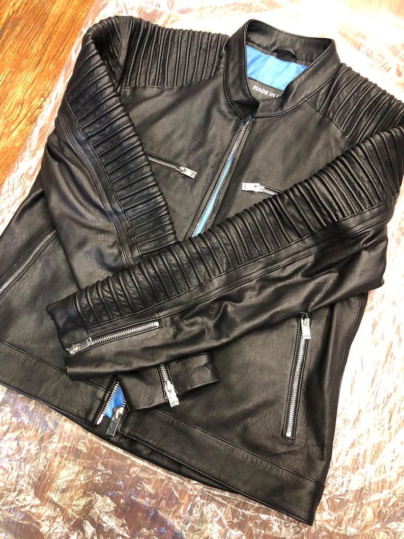 K&ouml;nnte beinhalten: Eine schwarze Lederjacke mit Stehkragen, leuchtend blauem Innenfutter und silberfarbenen Metallrei&szlig;verschl&uuml;ssen. Die &Auml;rmel und Schultern sind mit horizontalen Rippeneins&auml;tzen versehen und der Text MADE IN ist sichtbar.