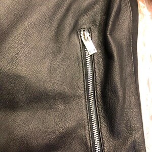 K&ouml;nnte beinhalten: Eine schwarze Lederjacke mit strukturierter Oberfl&auml;che und einer vertikalen Rei&szlig;verschlusstasche. Die Tasche verf&uuml;gt &uuml;ber einen silberfarbenen Metallschieber und Z&auml;hne, die sich vom dunklen, genarbten Material des Kleidungsst&uuml;cks abheben.