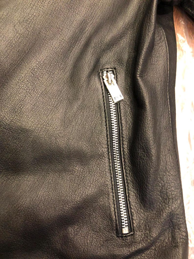 K&ouml;nnte beinhalten: Eine schwarze Lederjacke mit strukturierter Oberfl&auml;che und einer vertikalen Rei&szlig;verschlusstasche. Die Tasche verf&uuml;gt &uuml;ber einen silberfarbenen Metallschieber und Z&auml;hne, die sich vom dunklen, genarbten Material des Kleidungsst&uuml;cks abheben.